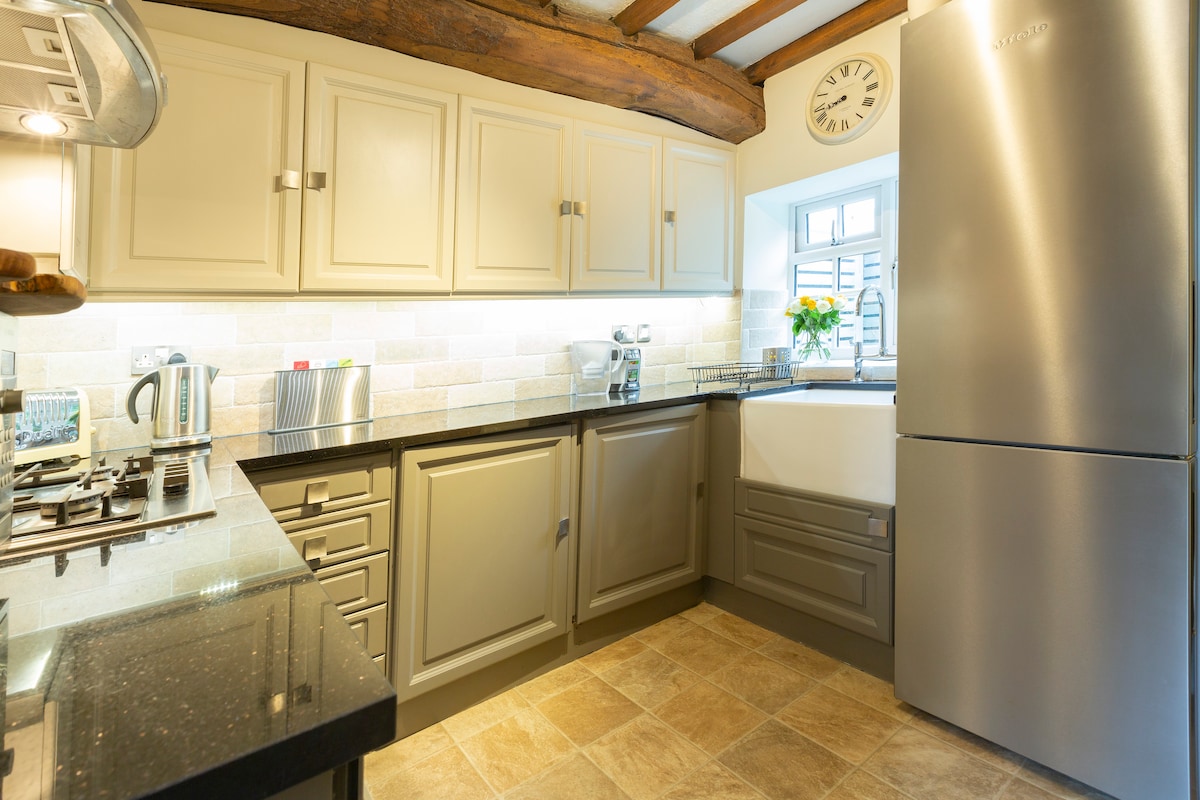 A modern kitchen is presented with stylish cabinetry, equipped with stainless steel appliances including a refrigerator and kettle. Natural light filters through a window, illuminating the neutral-toned walls and the dark countertop. A decorative clock adds a functional touch above.