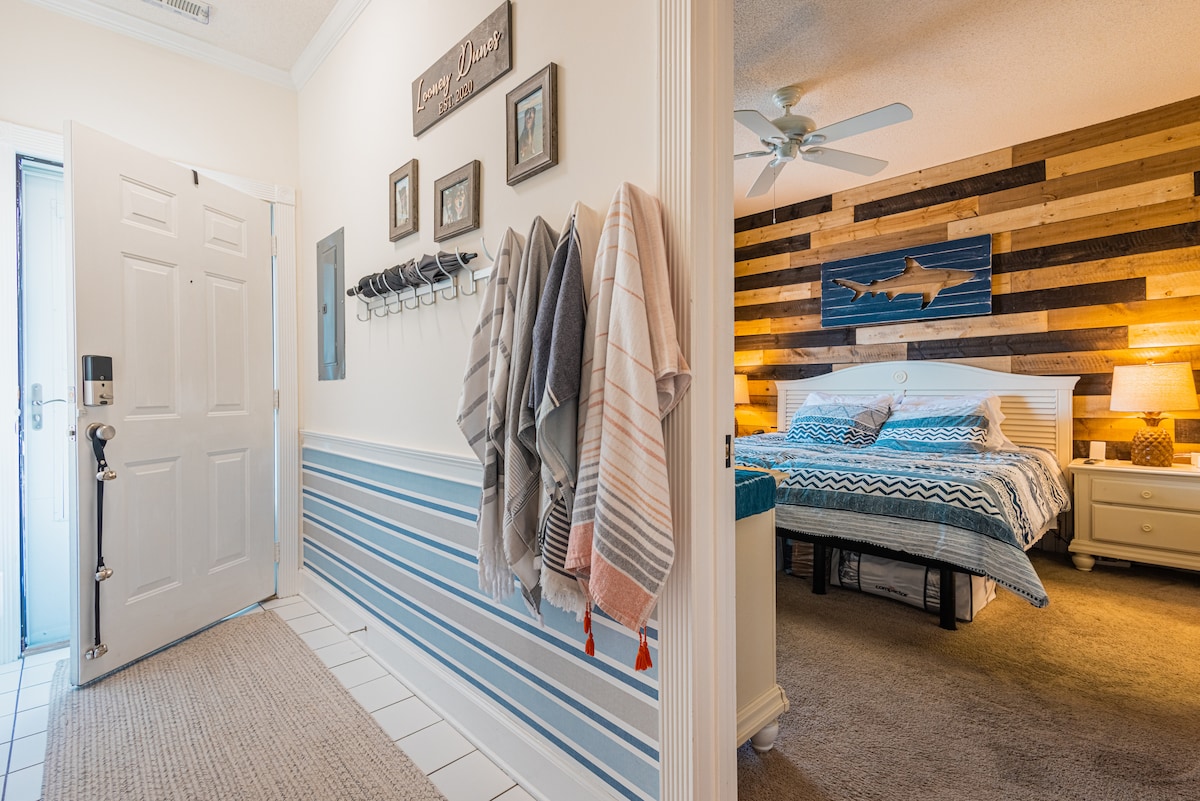 The entryway features a decorative wall with horizontal stripes and a rustic wooden accent wall behind the bed. Soft lighting illuminates the space, highlighting a neatly made bed with patterned linens. A set of hooks holds several blankets, adding to the welcoming atmosphere.