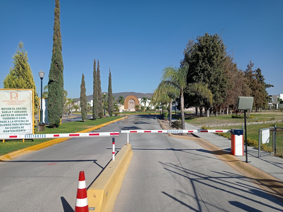 A gated entrance is presented, featuring a welcoming sign for visitors. A sleek security barrier is visible, with yellow curb and red-and-white striped markings. Lush trees line the street, under a clear blue sky, suggesting an organized and tranquil environment.