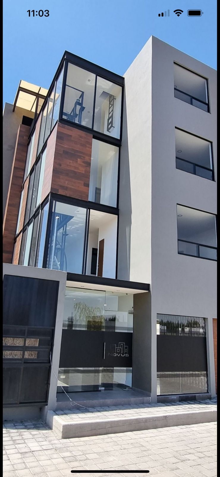 A modern multi-story building showcases a contemporary facade featuring large windows that invite natural light. The entrance is distinguished by a stylish design with glass and wood elements. A secure parking area is visible to the left, enhancing accessibility for guests.