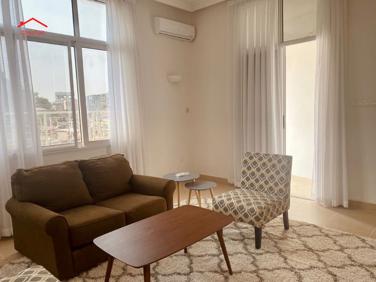 A cozy living room features a brown sofa and a patterned armchair, accompanied by a wooden coffee table. Large windows allow natural light to fill the space, with sheer white curtains gently flowing. A neutral rug adds warmth to the room, creating a comfortable atmosphere.
