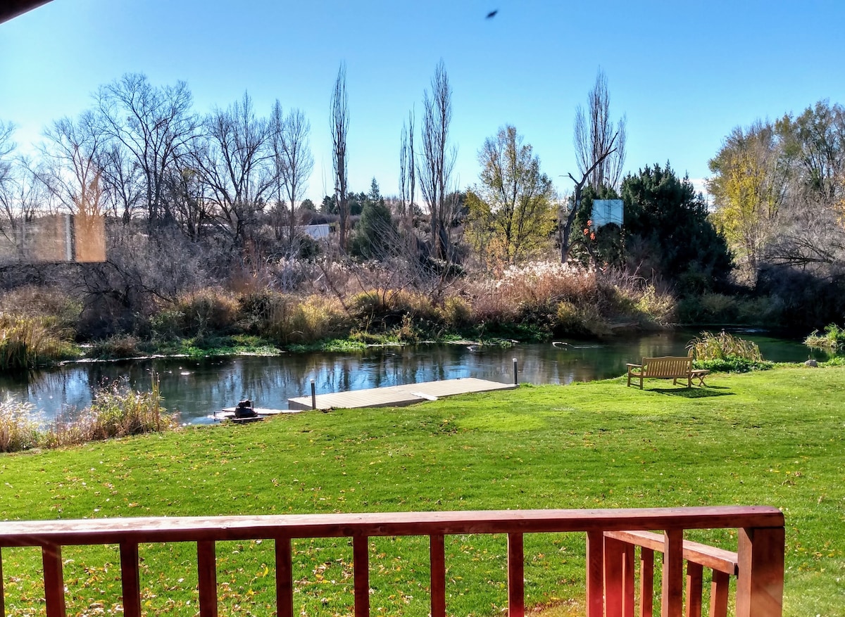 A view from the deck showcases lush green grass leading to the calm waters of Billingsley Creek. Trees line the creek's banks, and a wooden bench sits invitingly on the grass, providing a serene spot to enjoy the natural surroundings.