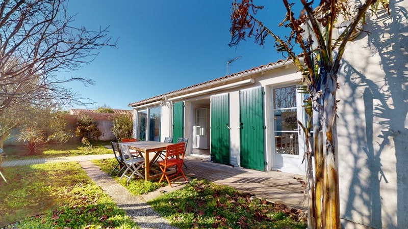 A single-story house is positioned in a garden setting, featuring green shutters and a wooden deck. A dining table with chairs occupies the outdoor area, surrounded by grass and various plants. Bright light illuminates the image, indicating clear weather.