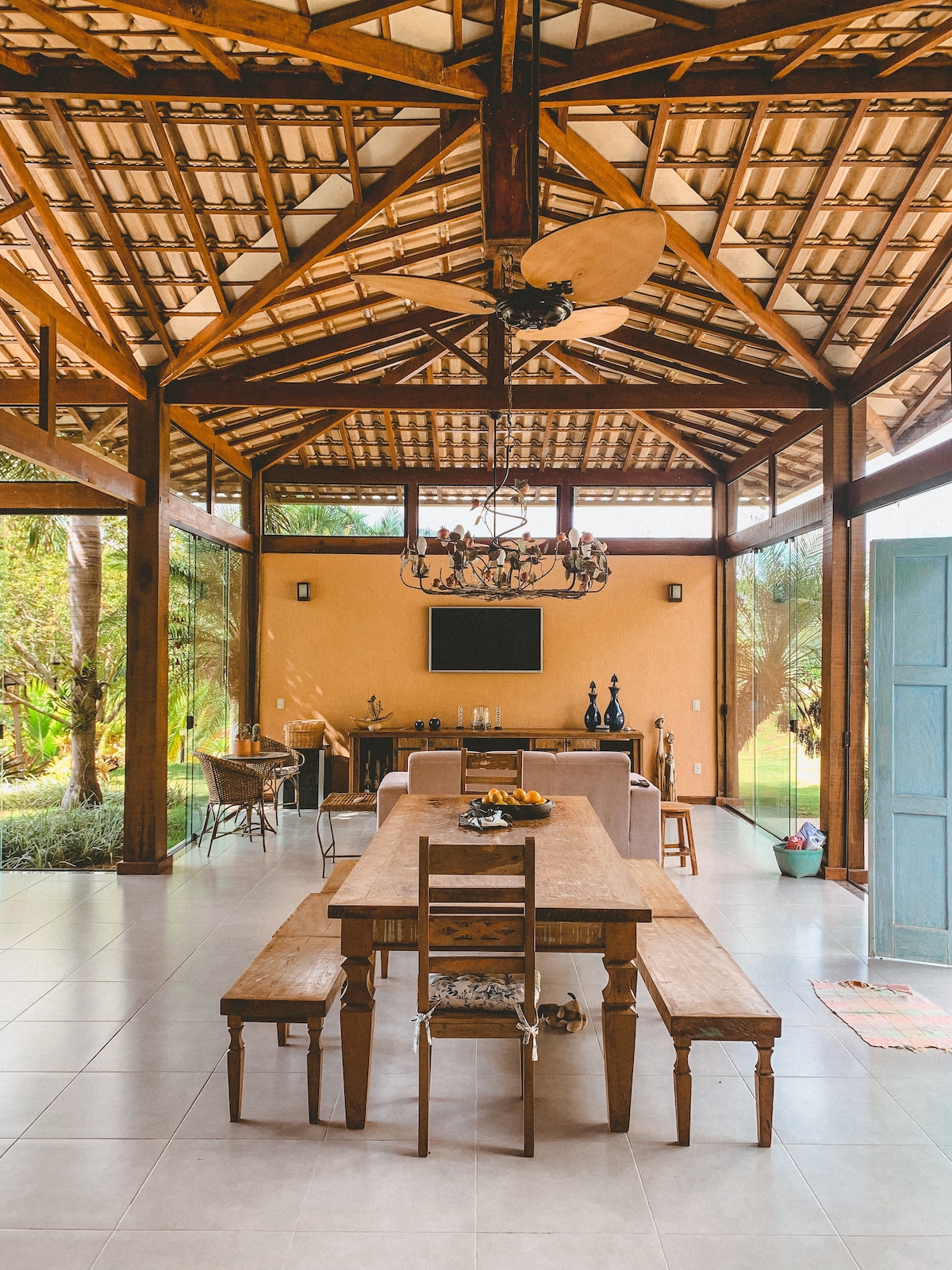 An open dining area features a long wooden table, surrounded by benches and chairs. The soaring ceiling is supported by wooden beams, with a ceiling fan above. Natural light flows in from large windows, showcasing lush greenery outside.