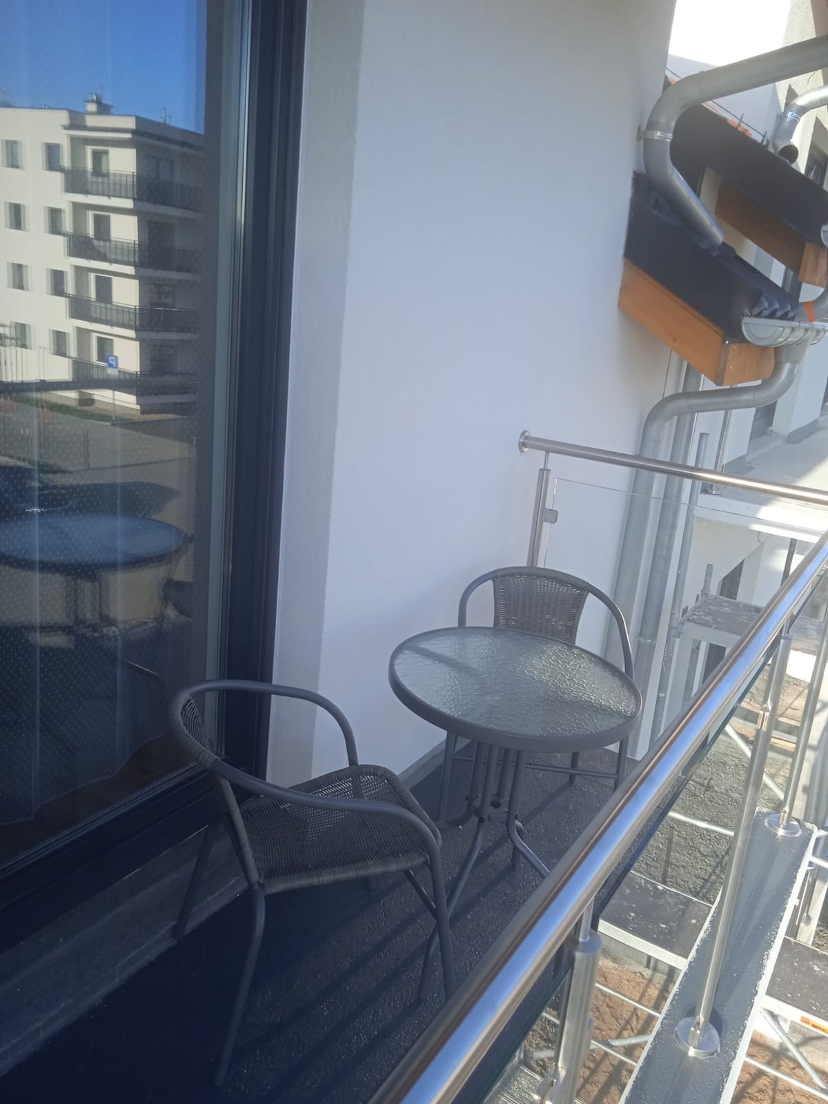 A small balcony features a round table and two woven chairs, providing a cozy outdoor space. The glass railing allows for clear views of the surroundings, with sunlight reflecting off the surfaces.