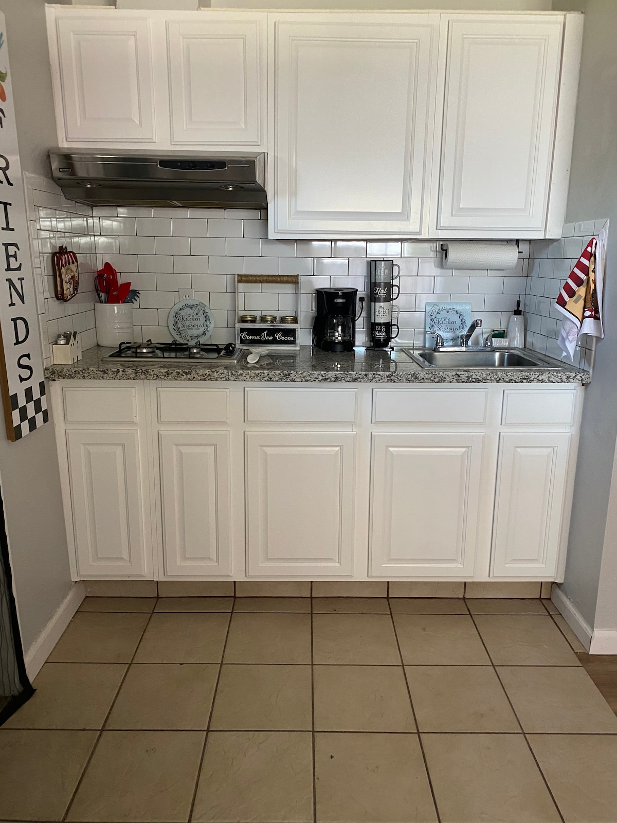 A compact kitchenette features white cabinetry and a granite countertop. A coffee maker sits beside a small stove and sink, with decorative dishware displayed above. The tiled floor adds to the space's welcoming feel, while natural light enhances the area.