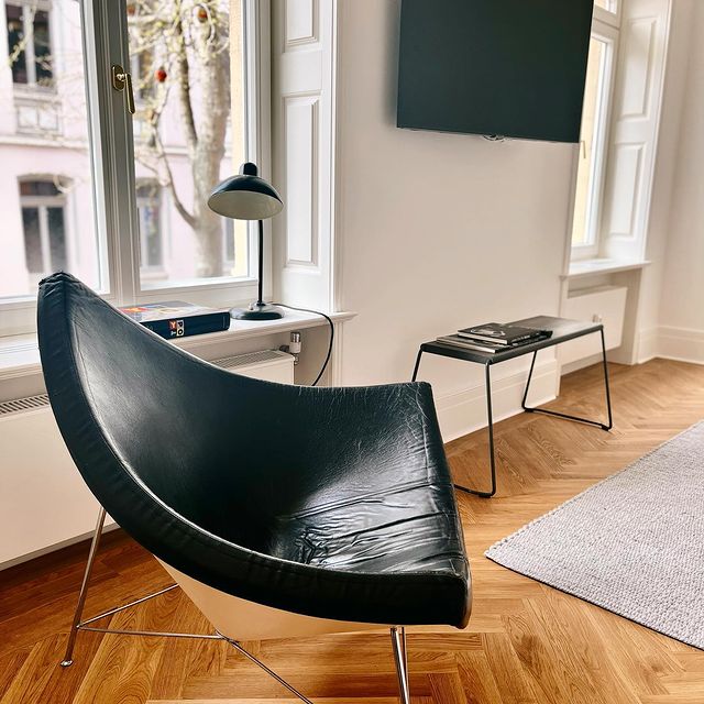 A sleek black chair is positioned near a large window, allowing ample natural light to enter the space. A minimalistic lamp stands on a side table beside it, with a rectangular coffee table featuring magazines placed on top. Light wooden flooring adds warmth to the area.