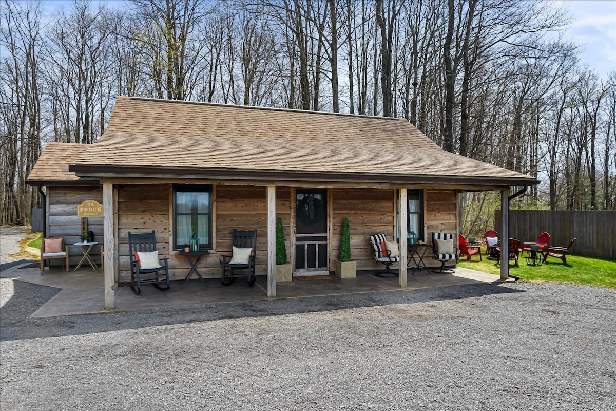 The cabin exterior features warm wood siding and a sloped roof. A covered porch is lined with rocking chairs and adorned with potted plants. In the yard, Adirondack chairs surround a fire pit, providing an inviting space for outdoor gatherings. The property is bordered by trees.
