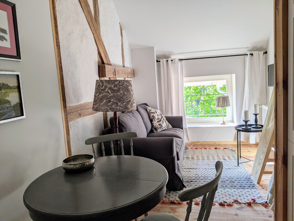 A cozy seating area includes a dark sofa adorned with decorative pillows, positioned opposite a small table and chairs. A lamp with a patterned shade provides soft lighting, while a window allows natural light to fill the space, revealing a view of greenery outside.