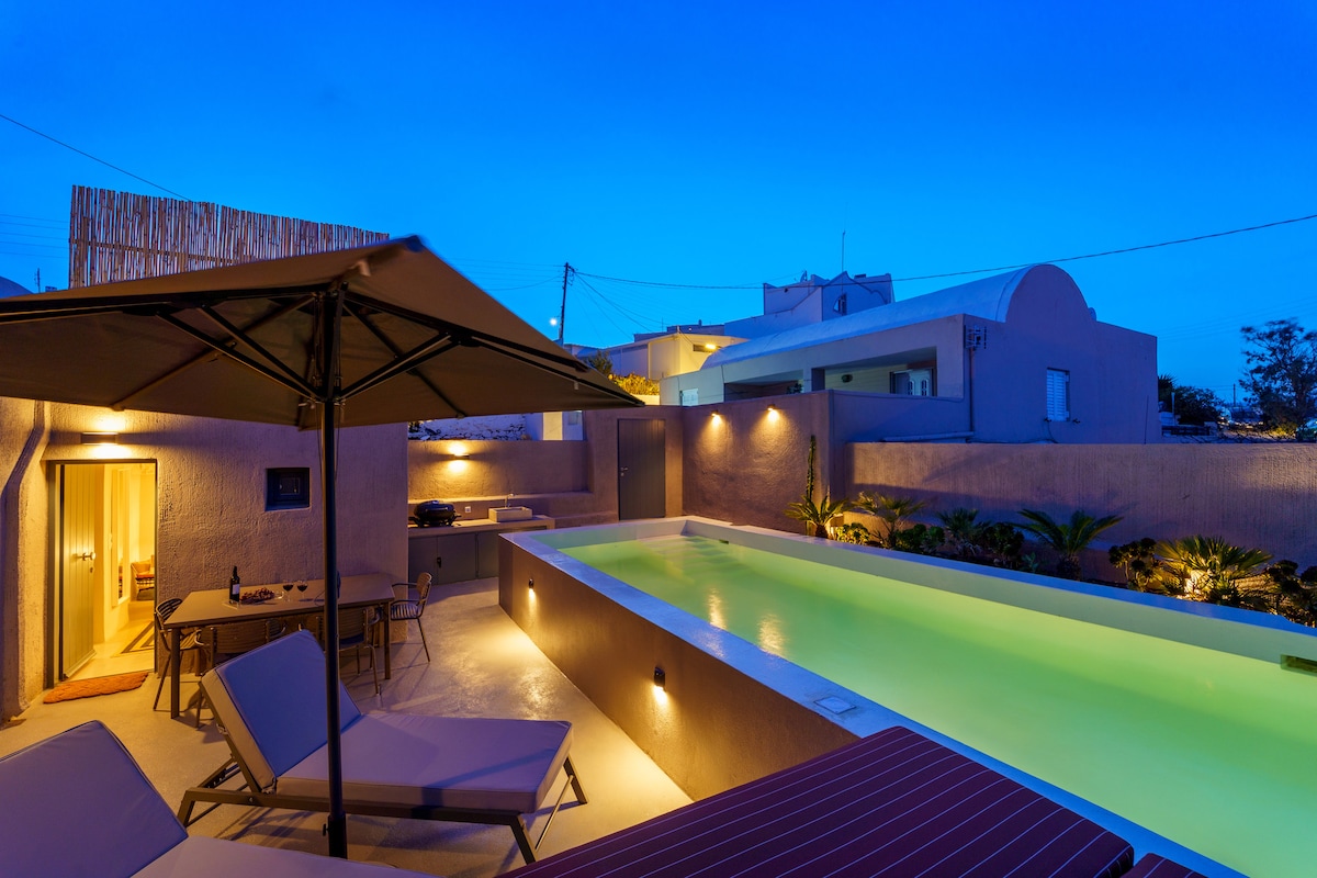 An outdoor area is illuminated by soft lights, featuring a private pool bordered by greenery. Sunbeds are arranged beside the pool, with a shaded seating area nearby. A doorway leads into the villa, blending indoor and outdoor experiences.