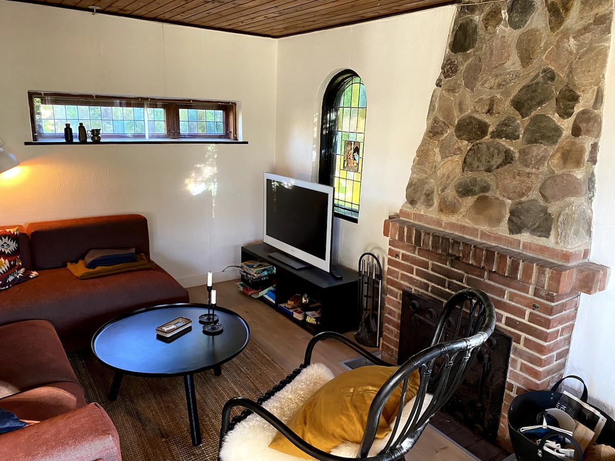 A cozy living area is arranged with a corner sofa and a black coffee table. A television rests opposite the seating, while a stone fireplace adds character. Light filters through a small window, creating a warm ambiance in the space.