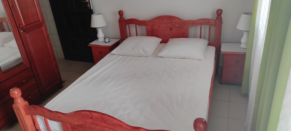 A wooden bed with a light-colored mattress is centered in the room. Two white pillows rest on top. Nightstands with lamps are positioned on each side, and a wardrobe is visible in the background, contributing to a functional and minimalist space.