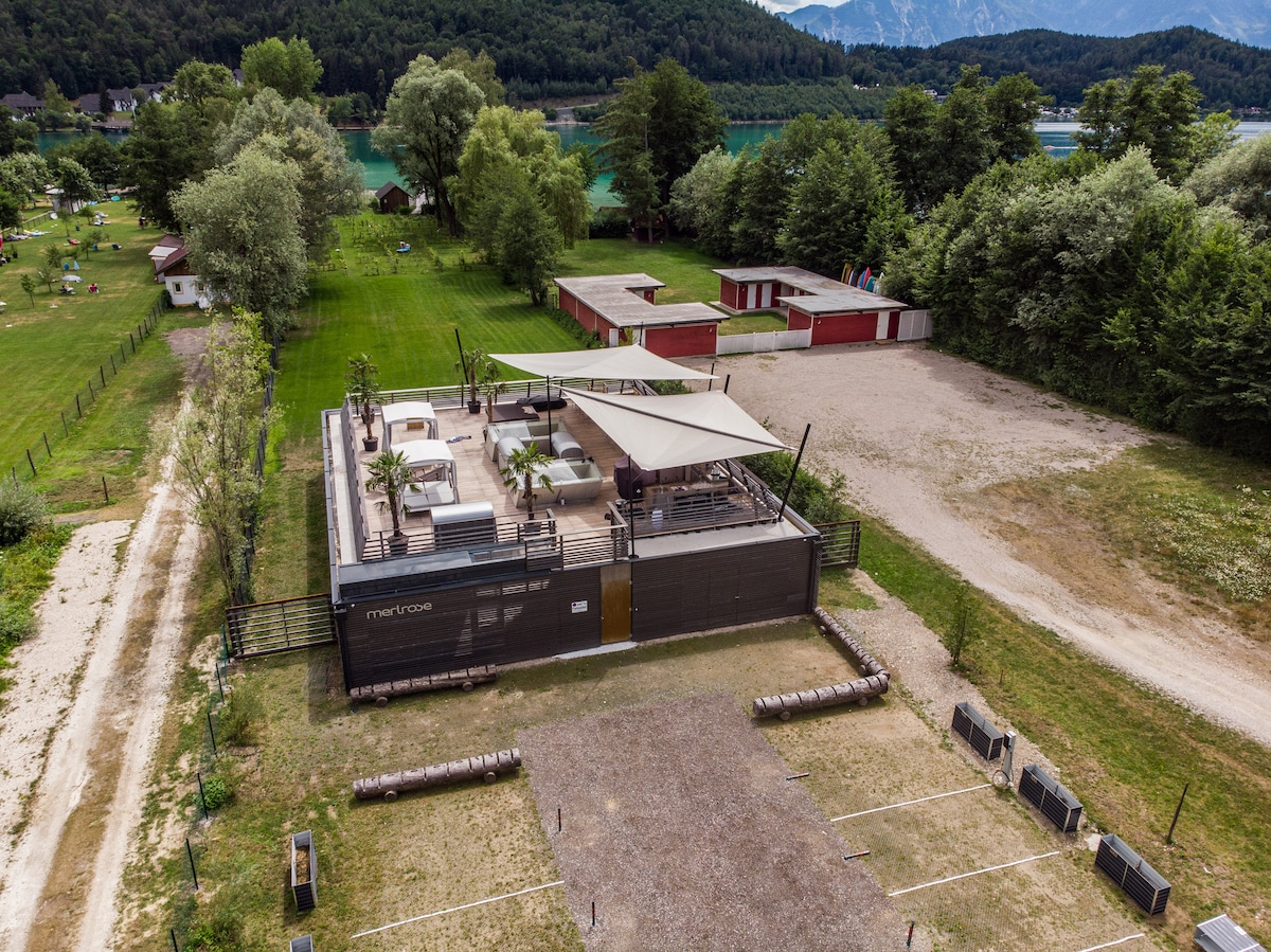 An aerial view of the Merlrose property, showcasing a spacious outdoor area with landscaped greenery and pathways. A modern structure with a shaded terrace is depicted, featuring seating areas. Surrounding amenities include parking spaces and storage units amid the natural surroundings near the Klopeiner See.