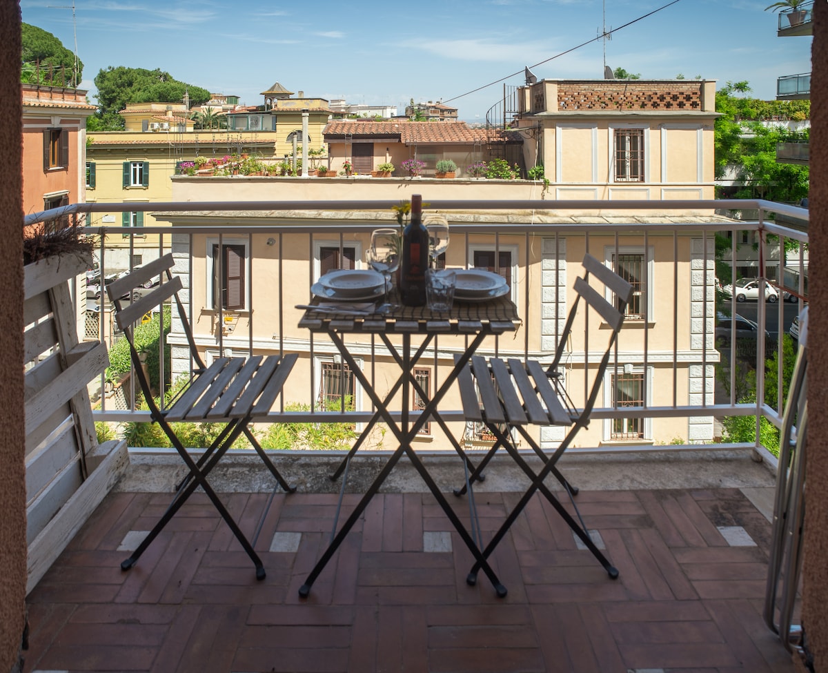 A small balcony features a round table set for two, accompanied by two wooden chairs with a modern design. The view includes neighboring buildings and greenery, suggesting a vibrant urban atmosphere. Sunlight illuminates the space, enhancing its open and inviting feel.