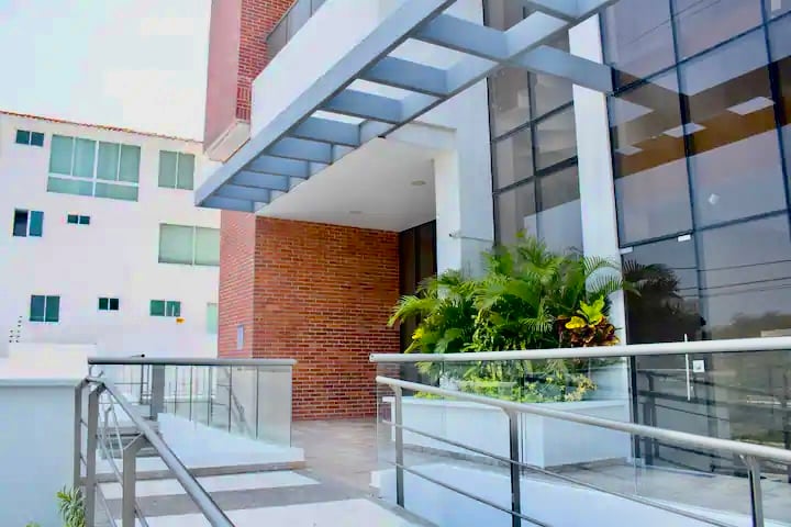 The exterior of the building is showcased, featuring a modern design with large glass panels. A ramp is visible, providing accessible entry. Lush greenery is present along the walkway, enhancing the inviting appearance of the entrance.