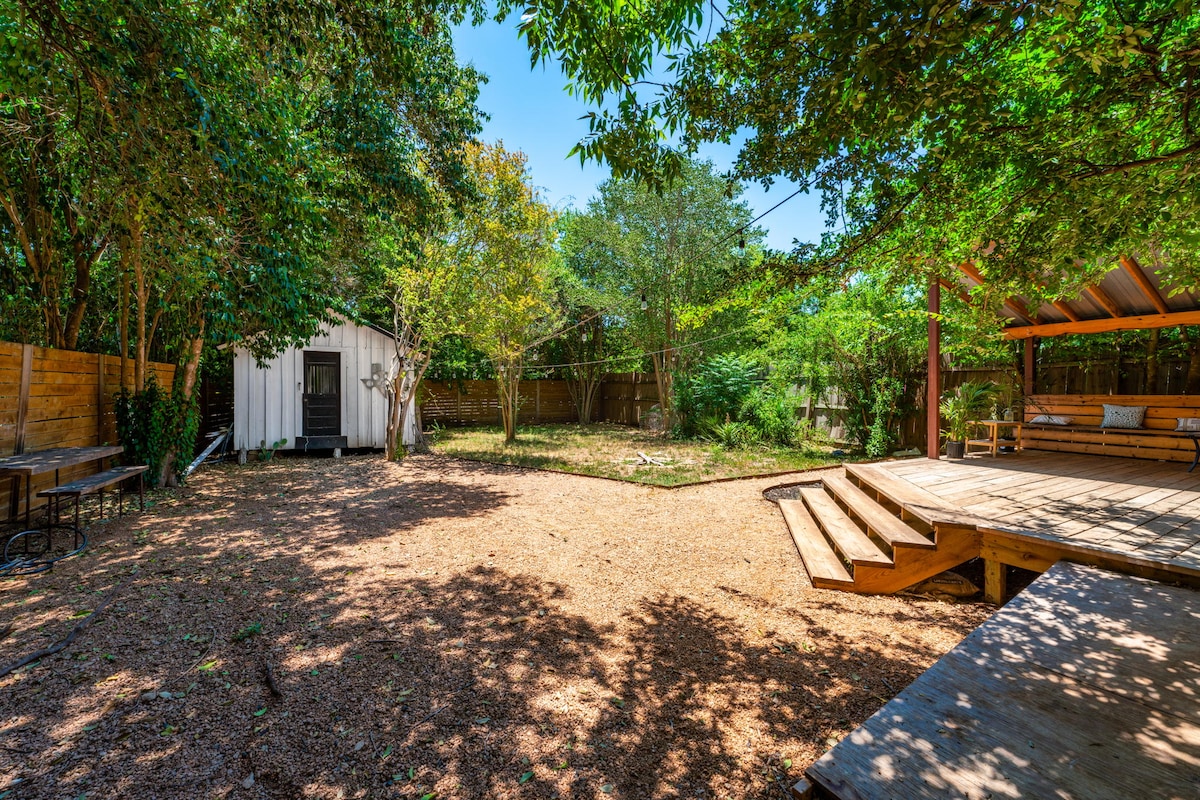 A spacious backyard is surrounded by lush trees, providing ample shade. A wooden deck extends into the space, and a white shed is positioned against the back fence. The ground is covered with soft tan mulch, creating a natural feel suitable for relaxation.