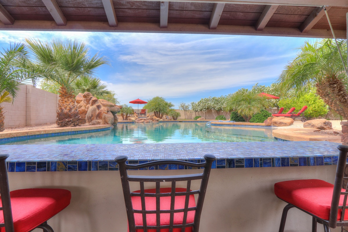 The view from the outdoor bar shows a large pool surrounded by palm trees and rock formations. Red umbrellas provide shade over lounge chairs nearby, while the sky features wispy clouds, enhancing the serene setting.
