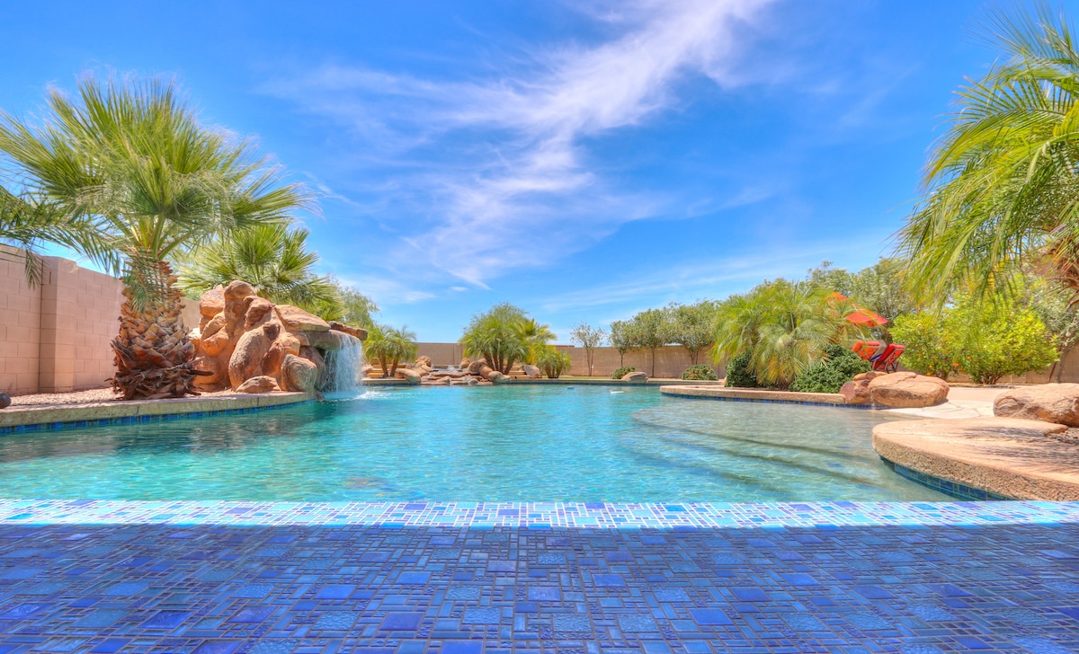 A large pool is surrounded by lush palm trees and rock formations, with a waterfall feature adding a serene touch. The inviting water reflects the clear blue sky, creating a tranquil outdoor retreat.