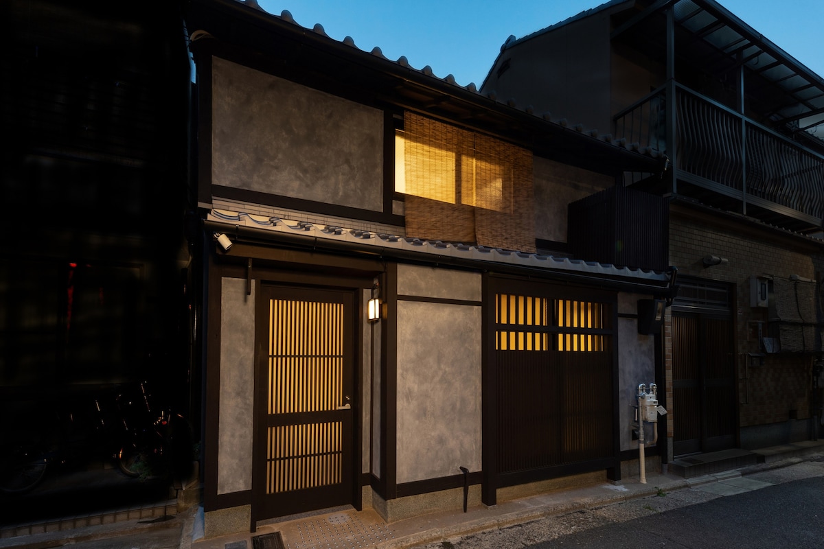 A traditional townhouse exterior features a blend of textured gray walls and wooden elements. Soft, warm light spills from the windows, highlighting the intricate wooden slat design. The entrance is framed by a welcoming door, creating an inviting appearance against the evening sky.