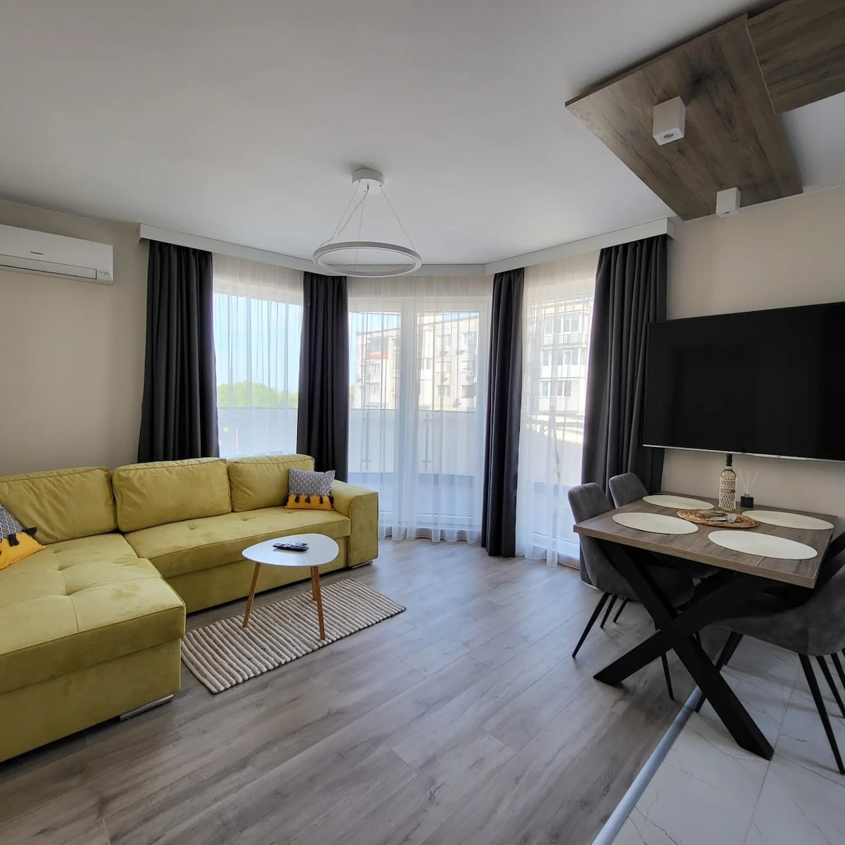 A modern living area features a comfortable yellow sectional couch, paired with a round table and four chairs. Natural light fills the space through large windows, with sheer curtains allowing for privacy. An entertainment unit with a mounted television is positioned against a wall.