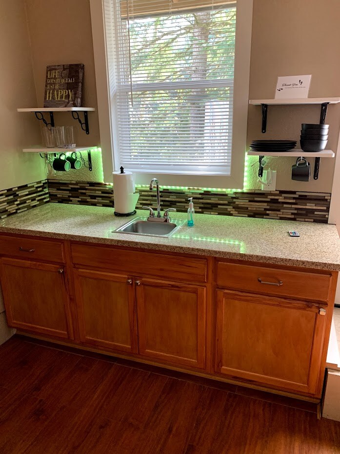 The kitchenette features wooden cabinetry and a granite countertop, equipped with a sink and essential cleaning supplies. Above, floating shelves hold ceramic dishes and utensils. Green accent lighting creates a welcoming ambiance, while a window provides natural light, illuminating the attractive backsplash.
