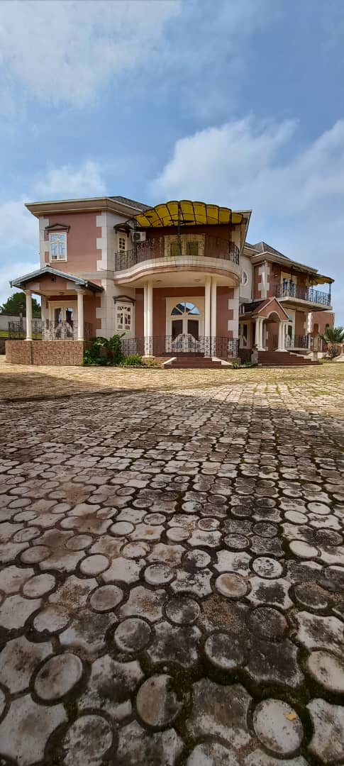 The exterior of the residence features a grand architectural design with a combination of stone and plaster. Large covered balconies are visible on the upper levels, and a spacious paved area leads up to the entrance. Lush greenery surrounds the property, enhancing its inviting presence.