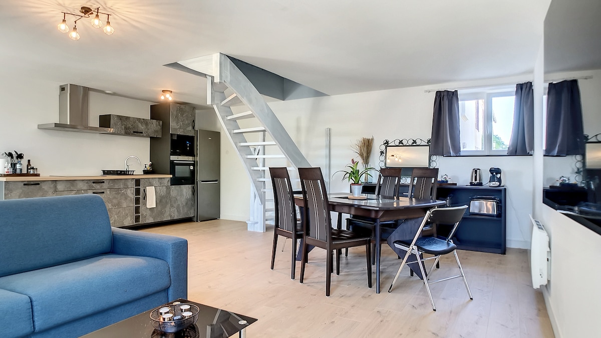 An open-plan living area features a comfortable blue sofa and a wooden dining table set for six. The kitchen is equipped with modern appliances, and a staircase leads to the upper level. Large windows allow natural light to brighten the space.