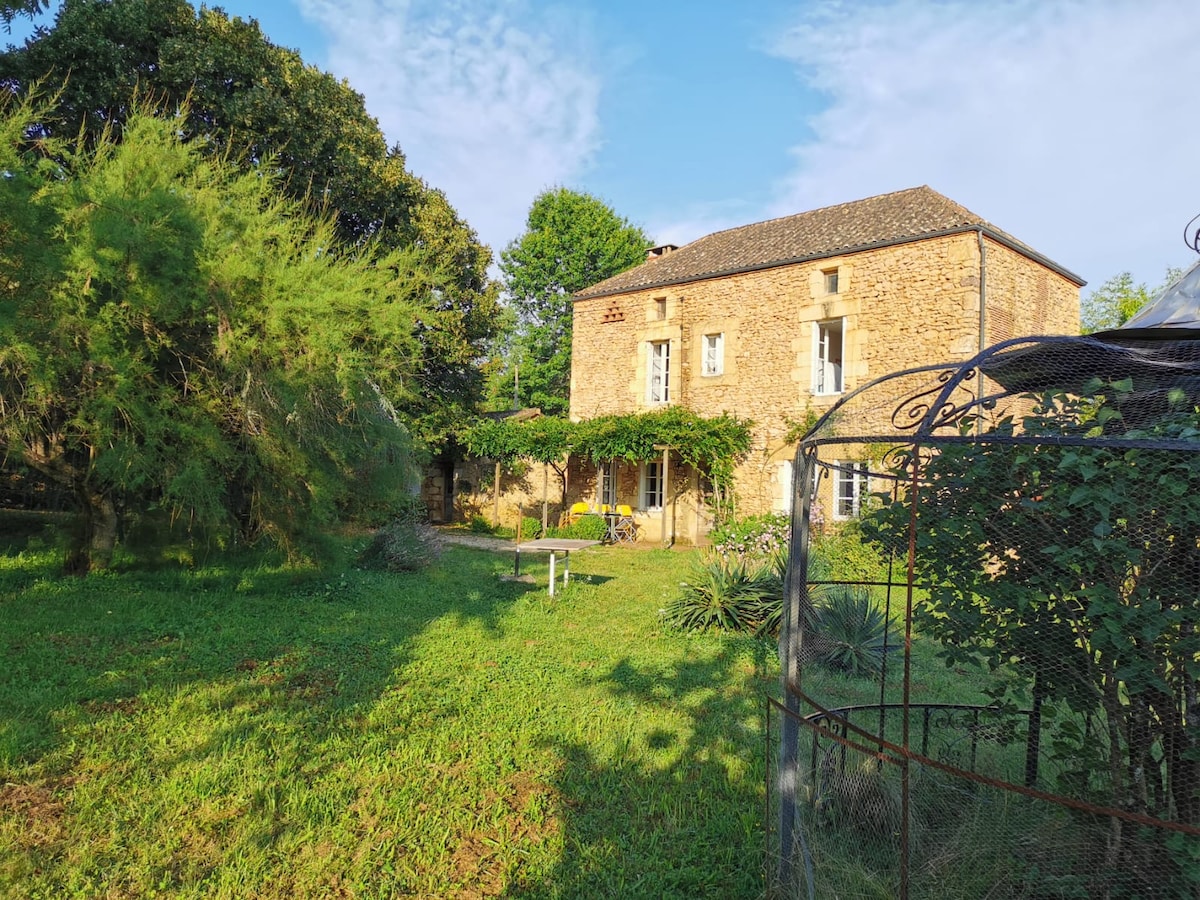 A stone house is set amidst a grassy area, surrounded by lush greenery and trees. The building features several windows and a climbing plant across the wall. A quaint outdoor seating area is visible, inviting relaxation in a peaceful environment.
