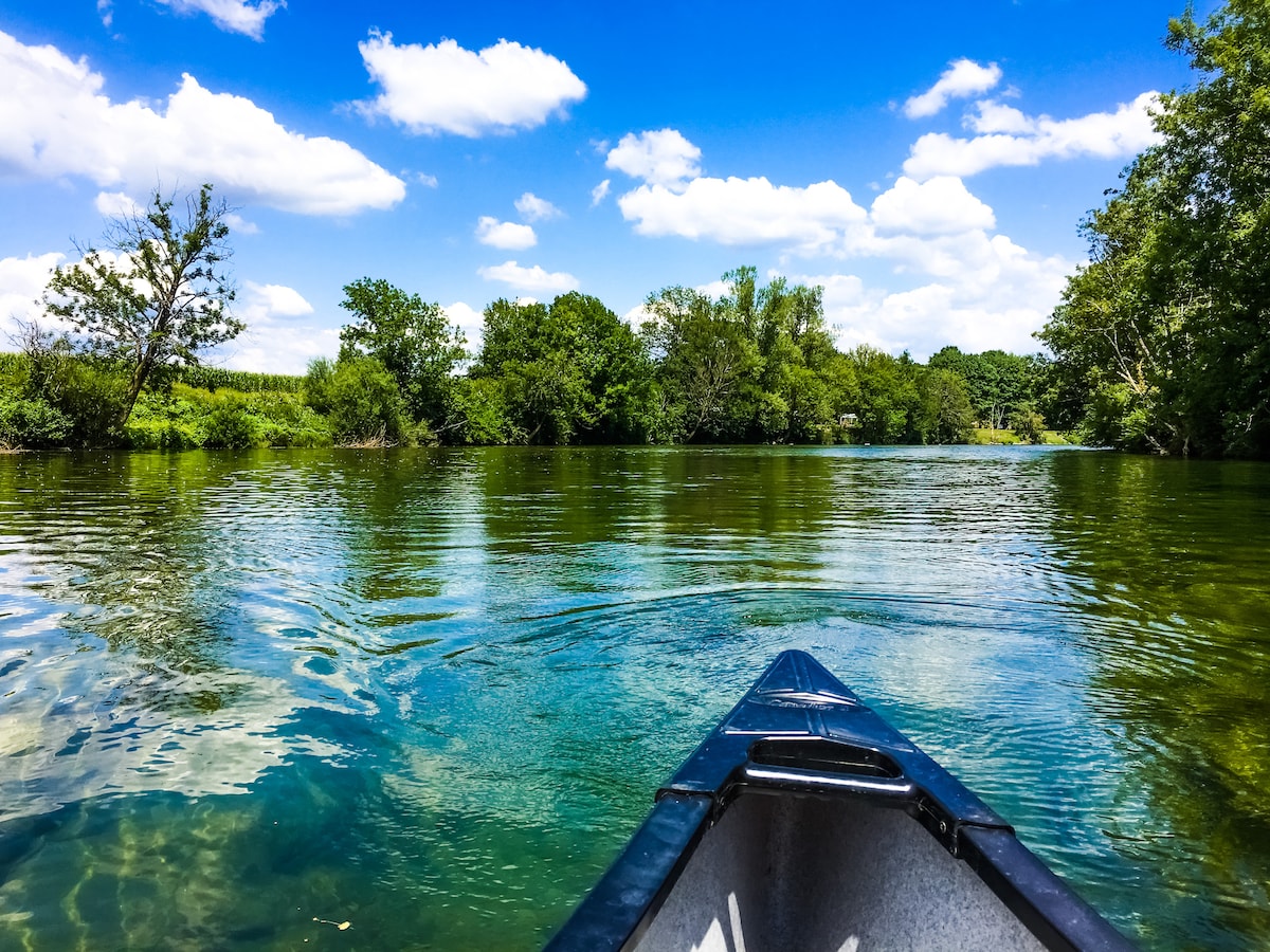 A view from a canoe looking down a serene river, with clear green waters reflecting the sky. Lush trees line the banks, and fluffy clouds are scattered across a bright blue sky, creating a peaceful natural setting.