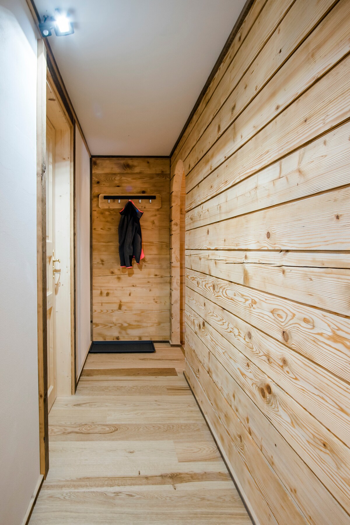 A well-lit hallway features wooden paneling that extends along the walls and a smooth wooden floor. A coat rack is mounted on the wall, and a small mat lies at the entrance. Bright lighting enhances the warm tones of the wood.