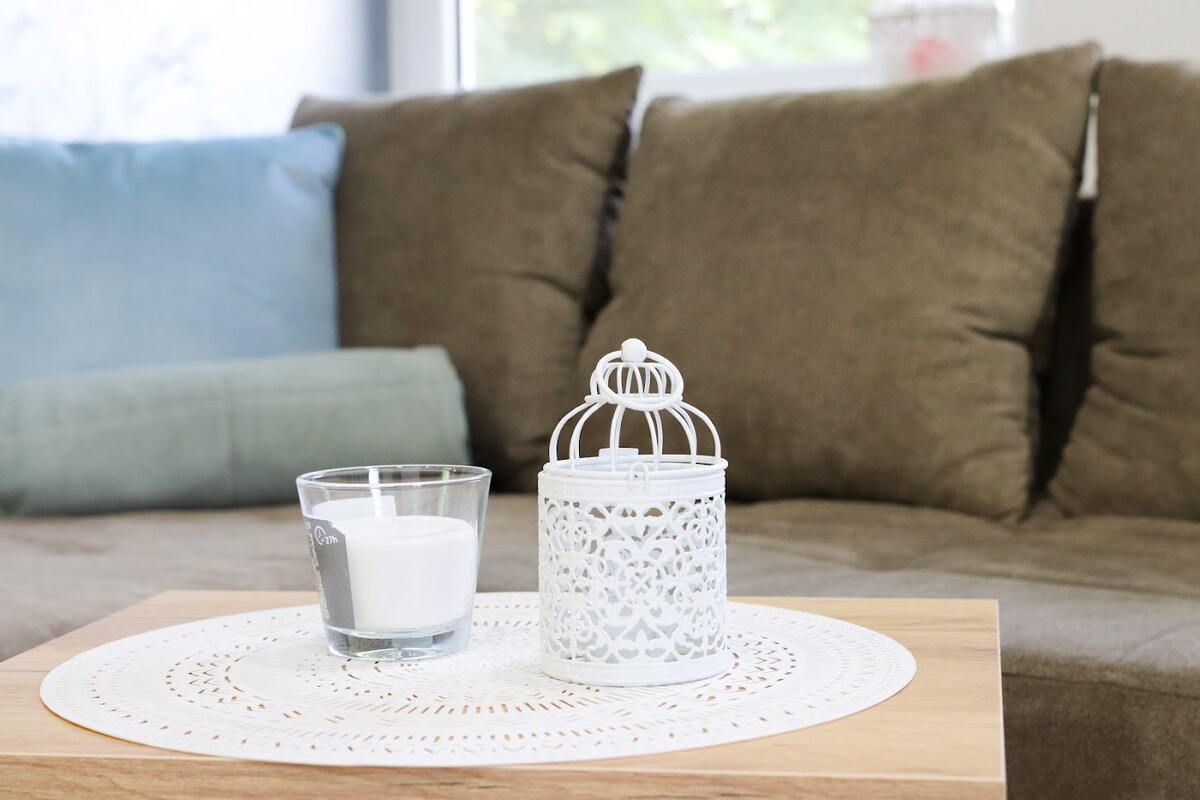 A cozy living area features a soft, brown sofa adorned with blue and green cushions. A wooden coffee table displays a glass candle and an intricately designed white lantern, positioned on an elegant round lace placemat.