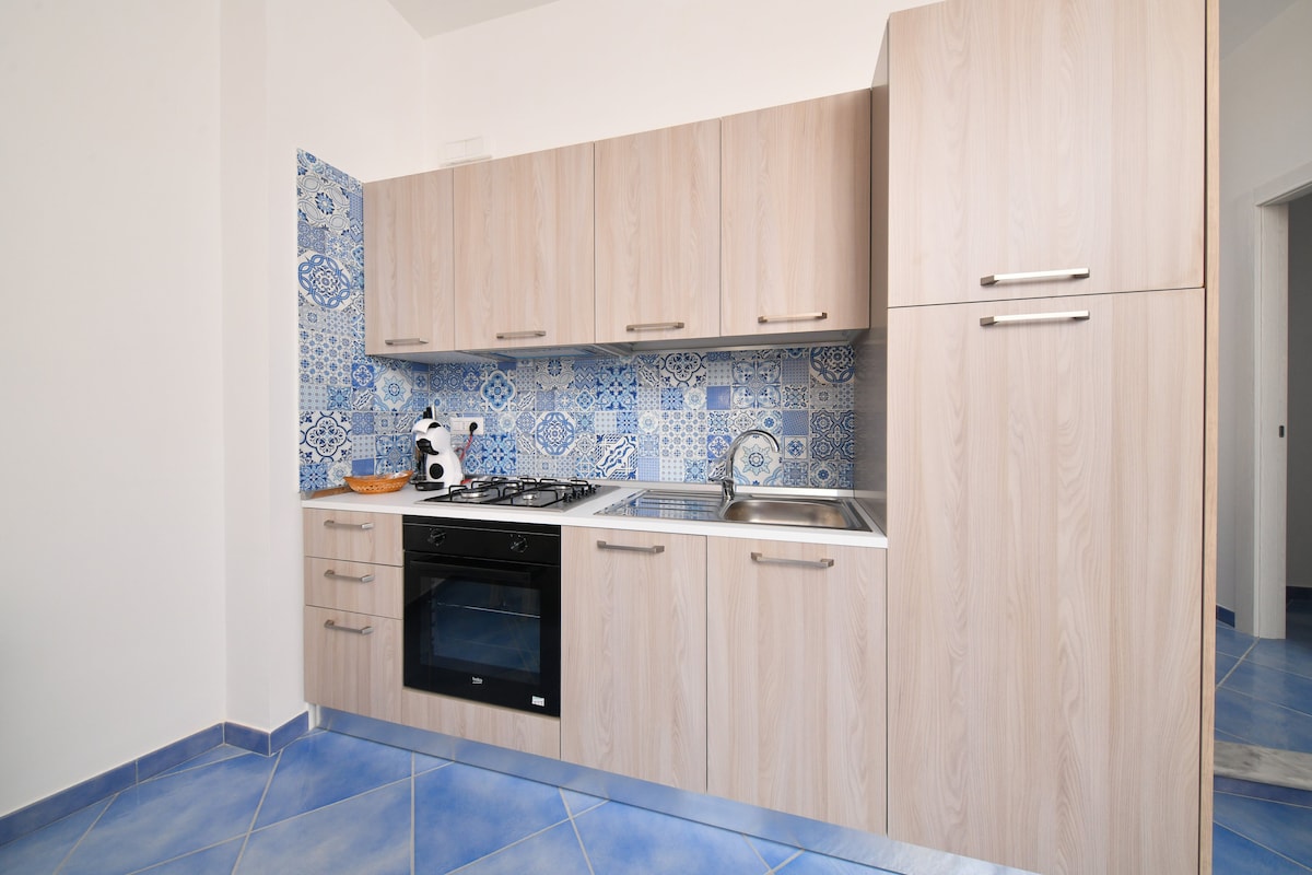 A modern kitchen area is featured with light wood cabinetry and a vibrant blue and white patterned backsplash. Stainless steel appliances, including an oven and cooktop, are positioned for convenience. The blue tiled flooring adds a cohesive color scheme to the space.