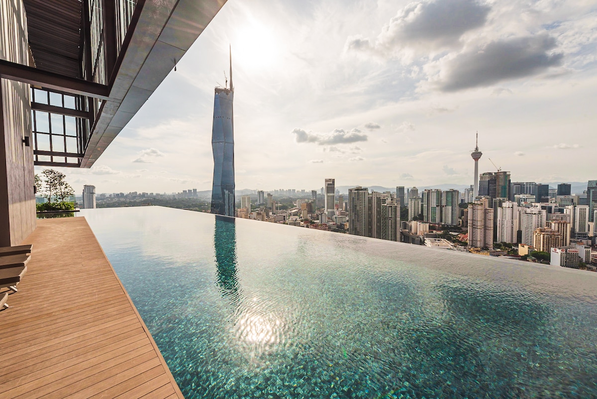 An infinity pool extends along a wooden deck, offering views of Kuala Lumpur's skyline. The water reflects the surroundings, with the PNB118 skyscraper prominently featured. Gentle waves ripple across the surface under a partly cloudy sky, enhancing the serene atmosphere of the space.