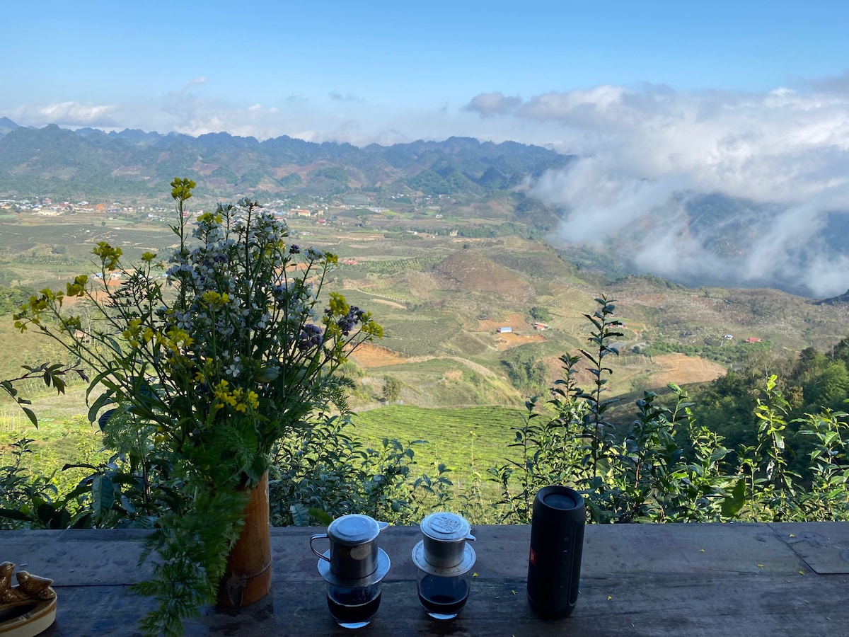 A table set against a scenic mountain backdrop features a vase of wildflowers and two cups. The expansive view reveals rolling hills and valleys, with clouds gracefully drifting across the landscape, creating a serene atmosphere.