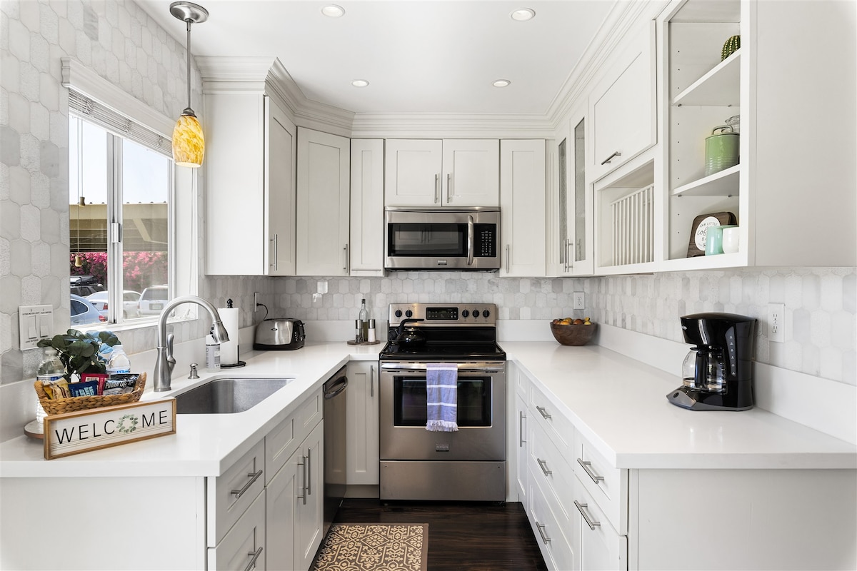 A modern kitchen is equipped with stainless steel appliances, including a microwave and coffee maker. White cabinetry and a gray tiled backsplash complement the spacious countertops. Natural light enters through a window, illuminating a decorative basket with a welcome sign.