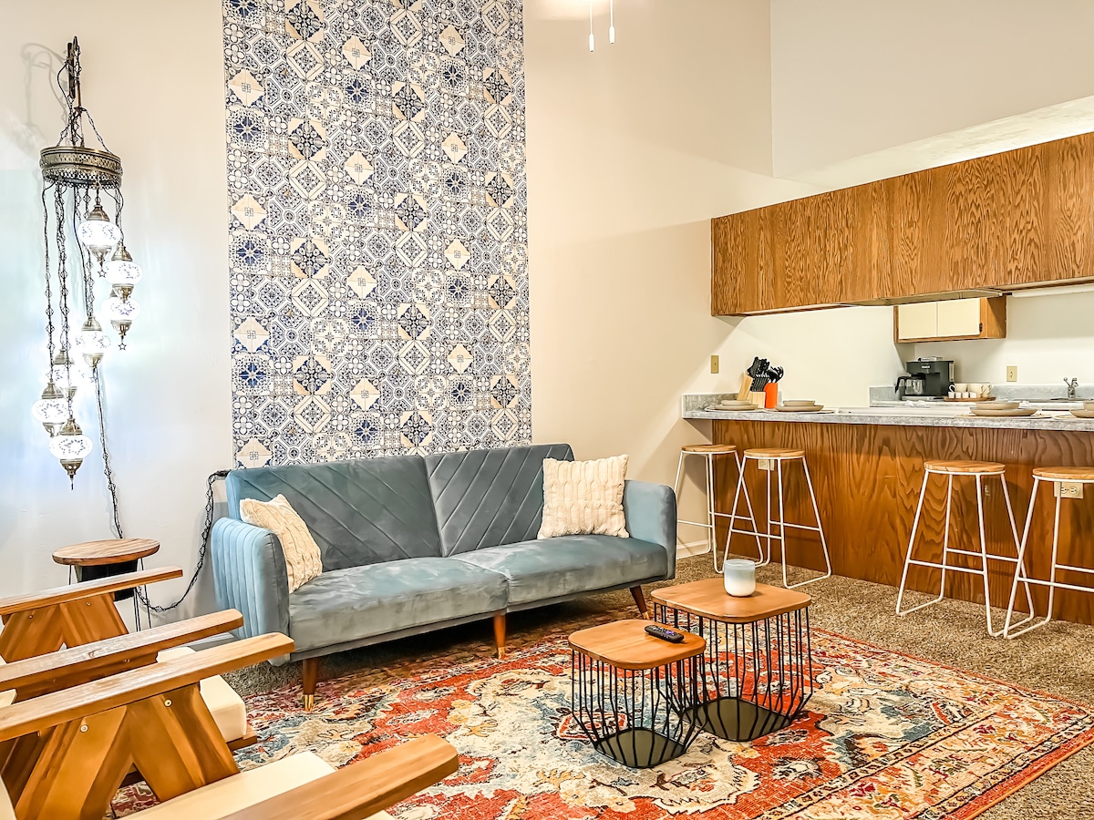 A spacious living area features a comfortable couch against a decorative blue and white patterned wall. Two wooden coffee tables sit on a colorful area rug, accompanied by modern stools at the kitchen counter visible in the background. Ample natural light brightens the space.