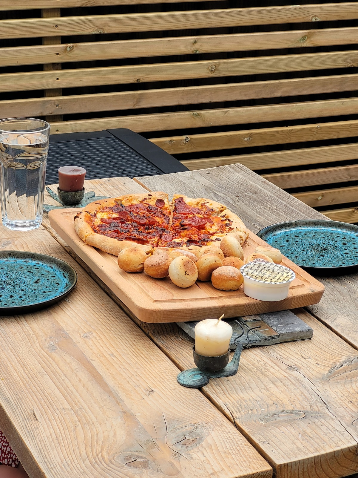 A wooden table is set with a variety of food items. A freshly baked pizza, topped with cheese and pepperoni, is presented on a wooden platter, accompanied by dough balls arranged nearby. Two blue plates are placed on the table, along with a glass of water and a candle.