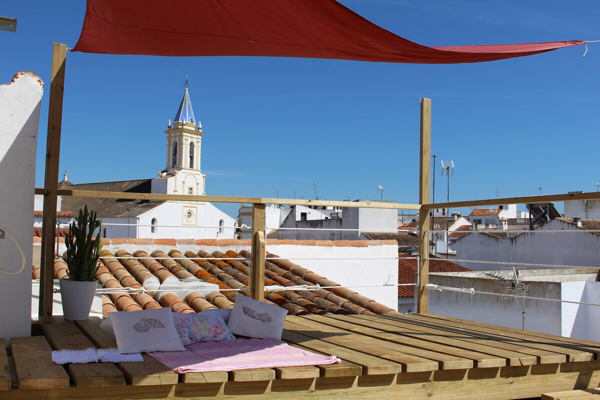 A wooden deck is shown on the rooftop, furnished with two cushions and a decorative throw. A potted plant enhances the space, with a bright blue sky in the background. A church steeple is visible among the whitewashed buildings of Cartaya.