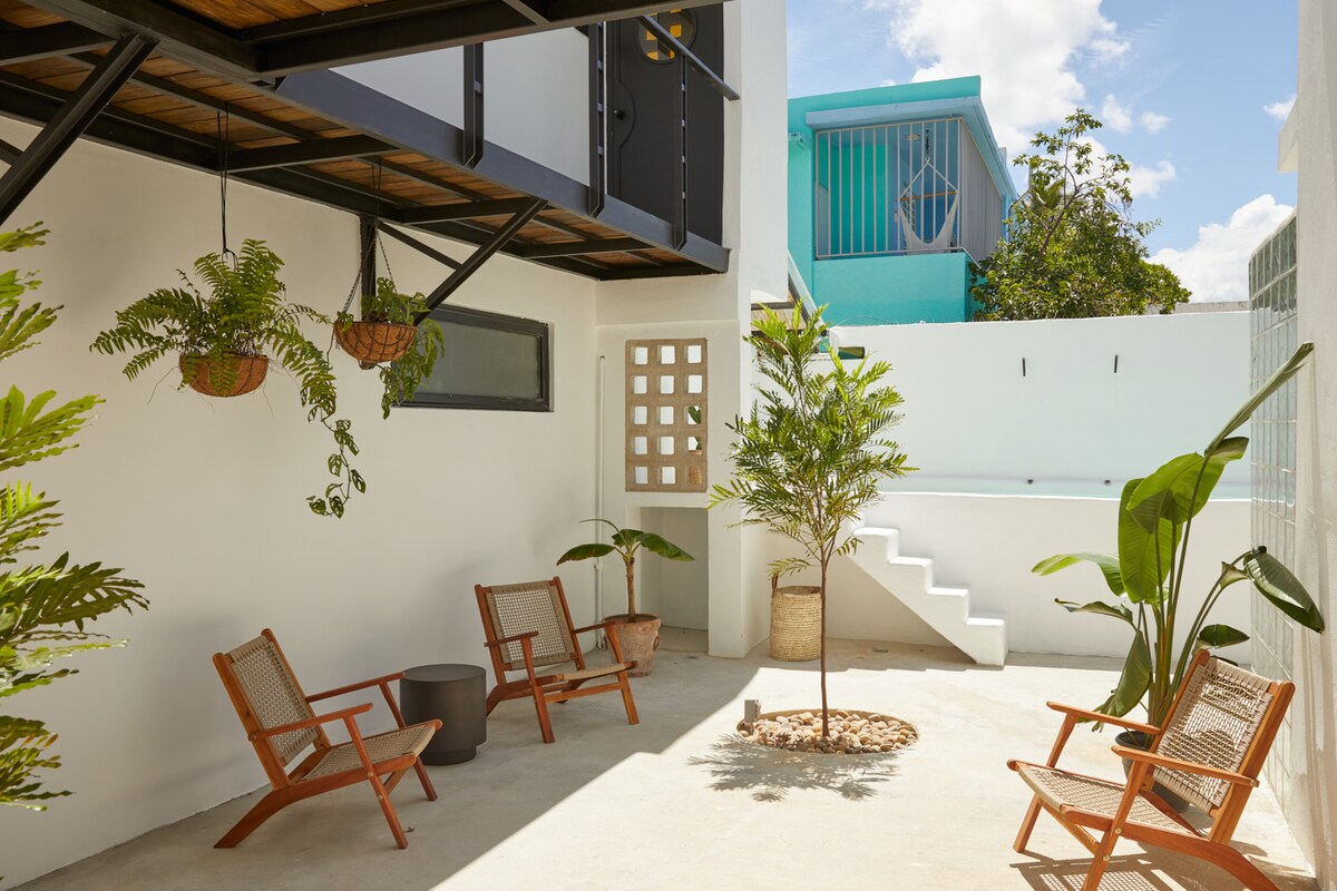 An inviting outdoor courtyard features four mid-century chairs arranged around a small table. Tropical plants are placed in decorative planters, while a small tree stands centrally. A staircase leads to a shared pool space, set against a backdrop of bright blue accents.