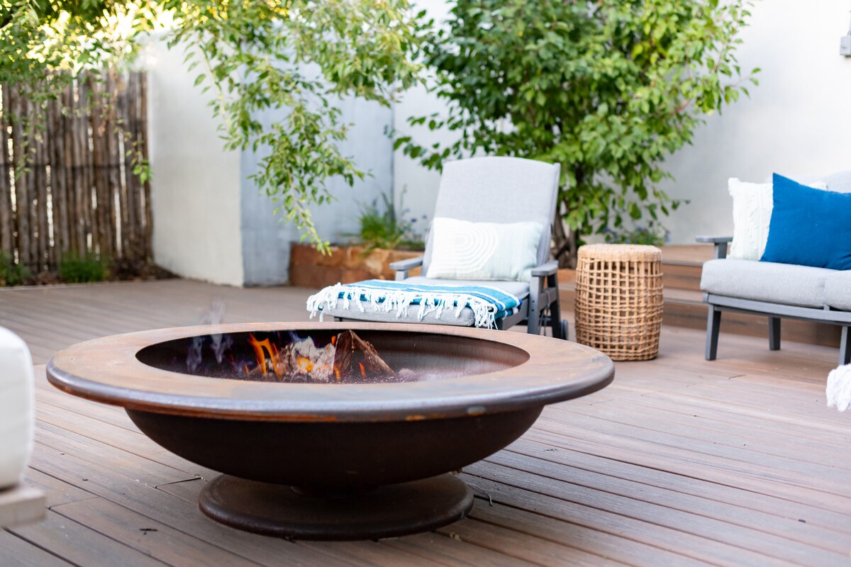 A cozy outdoor seating area features a fire pit in the center, surrounded by wooden decking. Two chairs are positioned nearby, one adorned with a blue cushion. Lush greenery adds privacy and a natural touch to the serene space.