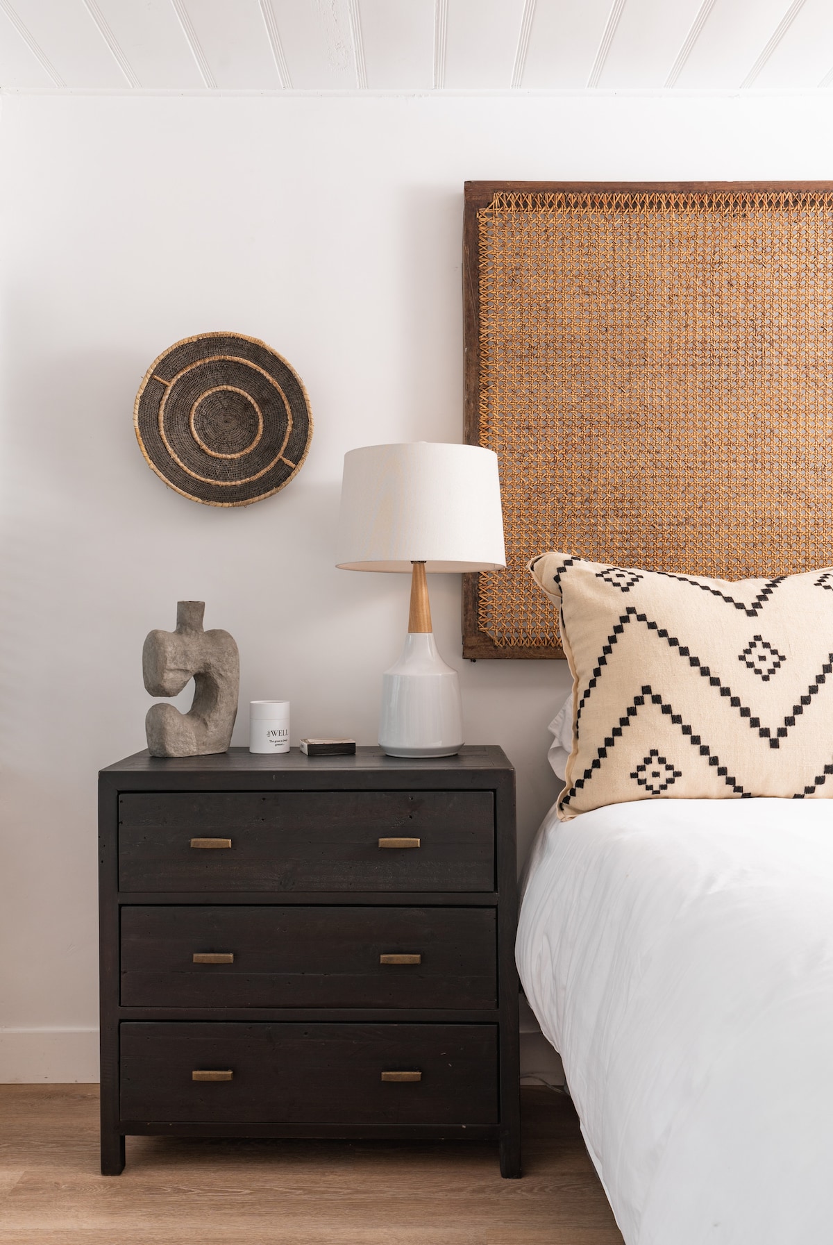 A bedside table features three drawers, topped with a decorative lamp and an abstract sculpture. A patterned pillow rests against the headboard, which is accented by a woven wall hanging, adding texture to the serene sleeping area.