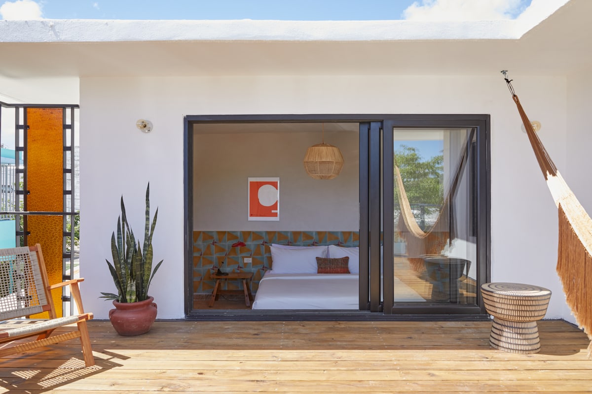 A spacious outdoor patio showcases a hammock and wooden lounge chair, framed by potted plants. Large glass doors provide a glimpse into a bedroom featuring a vibrant accent wall and a warm color palette. Natural light fills the space, enhancing the inviting atmosphere.