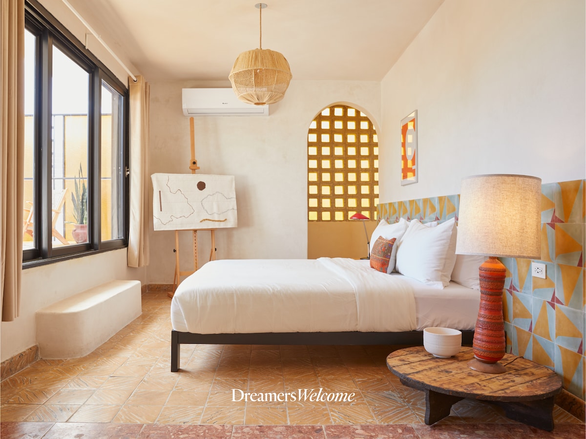 A serene bedroom features a spacious bed with crisp white linens. A decorative light fixture hangs above and a wooden side table stands beside the bed. Large windows allow natural light, complemented by geometric tiles on the headboard and warm terracotta flooring.