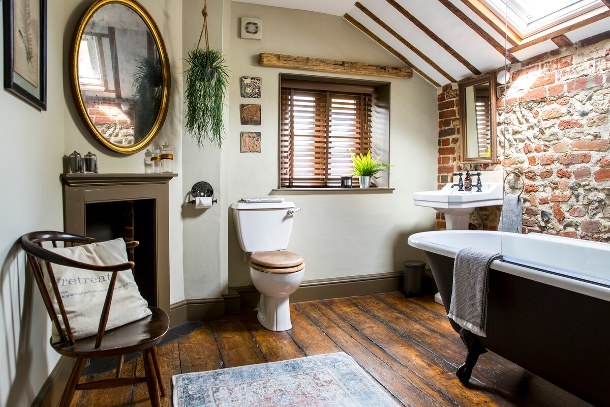 A bathroom features a freestanding bathtub beside a window with wooden shutters. Wood flooring complements exposed brick walls and decorative elements. A round mirror is mounted above a dark wood shelf, and a towel hangs on a rail near the stylish toilet. A soothing plant enhances the space.