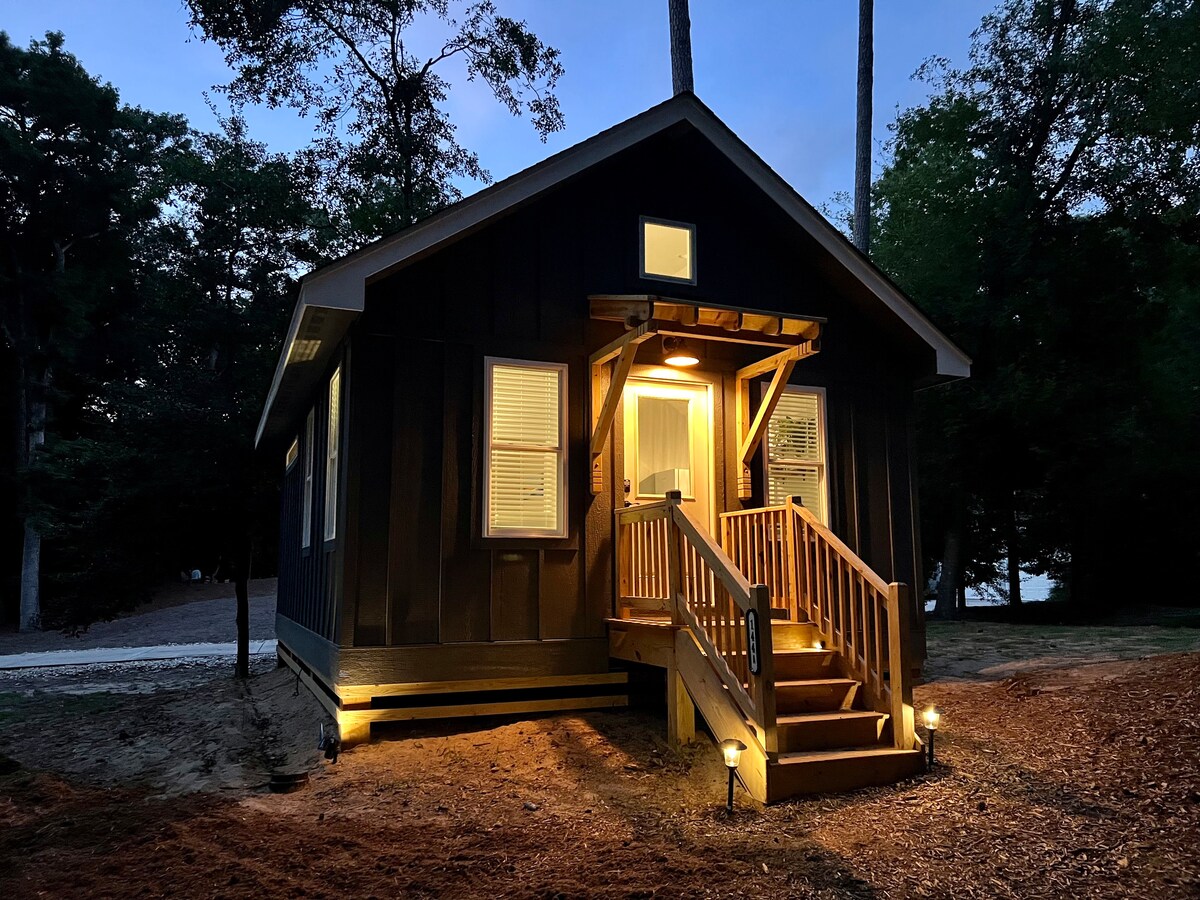 The exterior of a cozy cottage is illuminated by soft outdoor lighting, highlighting the rustic wooden stairs leading to the front door. Large windows frame the entrance, while surrounding trees add a touch of nature to the peaceful setting.