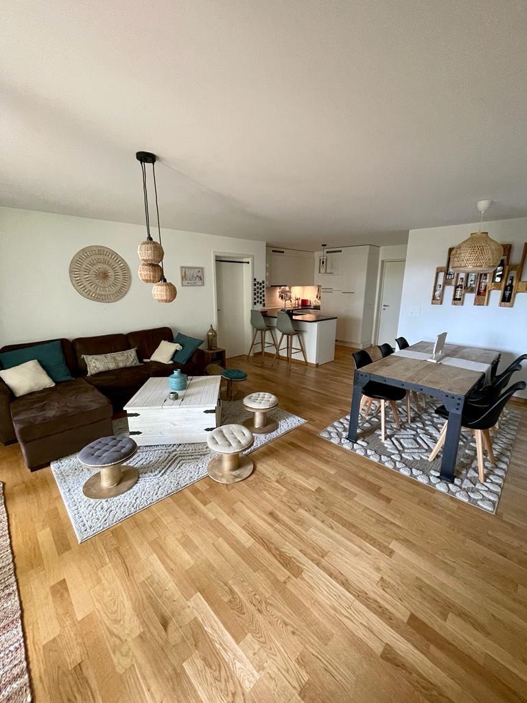 A spacious living area features a dark brown sofa accompanied by decorative cushions. A light wood dining table is surrounded by black chairs, with stylish lighting fixtures hanging overhead. The floor showcases warm wooden tones, complemented by two textured area rugs in neutral shades.