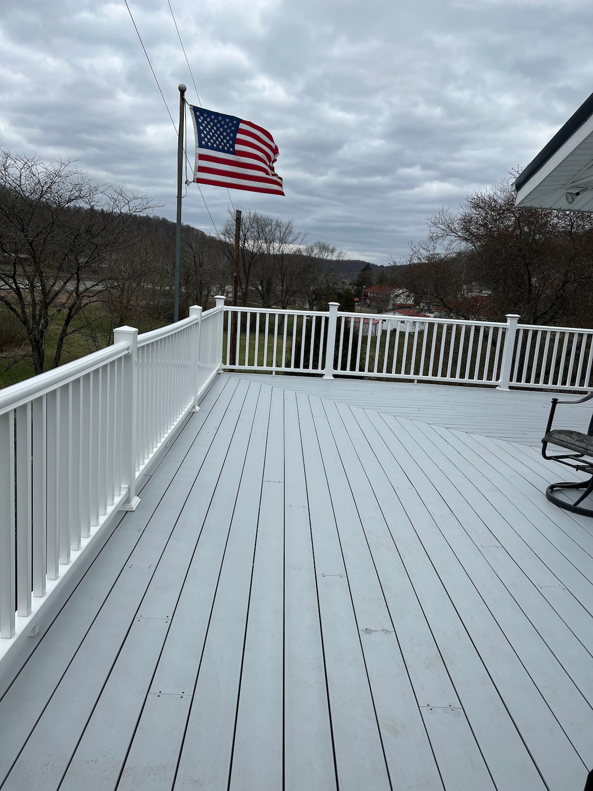 A spacious porch is shown, featuring light gray flooring and a white railing. An American flag waves gently in the breeze, with a scenic view of the surrounding landscape visible in the distance. A rocking chair is positioned to offer a cozy spot for relaxation.