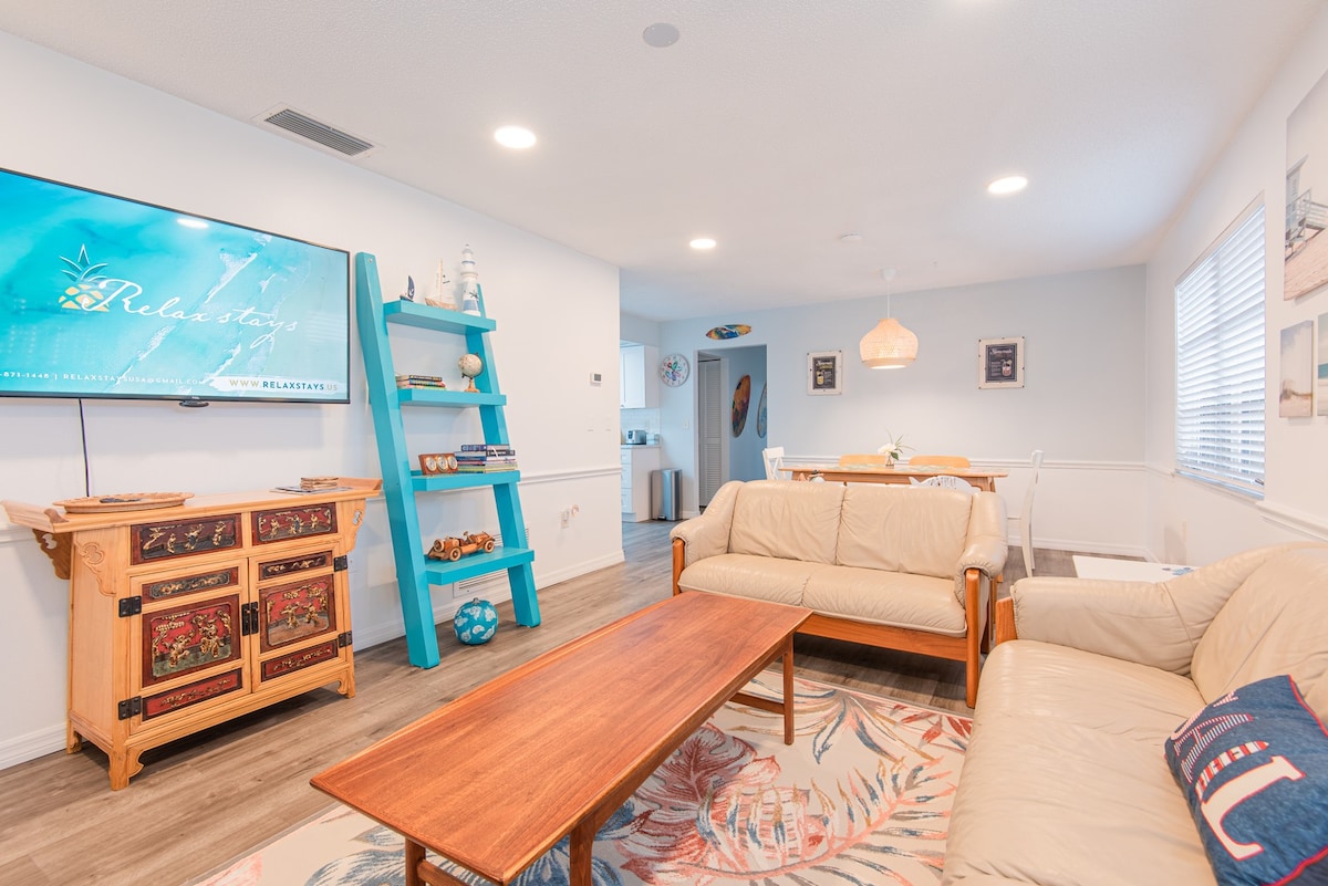 An open concept living area is captured, featuring a wooden coffee table centered between a light-colored sofa and loveseat. A blue bookshelf holds decorative items. A television is mounted on the wall, while the dining area is subtly visible in the background.