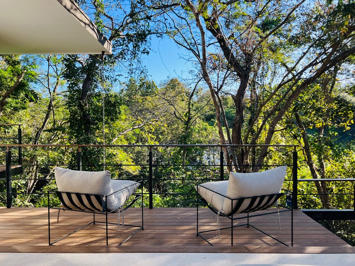 A wooden deck is shown with two comfortable chairs arranged facing a view of lush greenery and trees. The space offers a sense of openness, framed by a sleek railing that complements the natural surroundings.