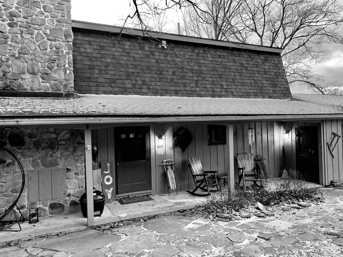 The exterior of a charming old cottage is displayed, featuring a stone wall and wooden siding. Rocking chairs are positioned on the porch, and a wreath adorns the dark front door. A gravel pathway leads to the entrance, surrounded by natural landscaping.